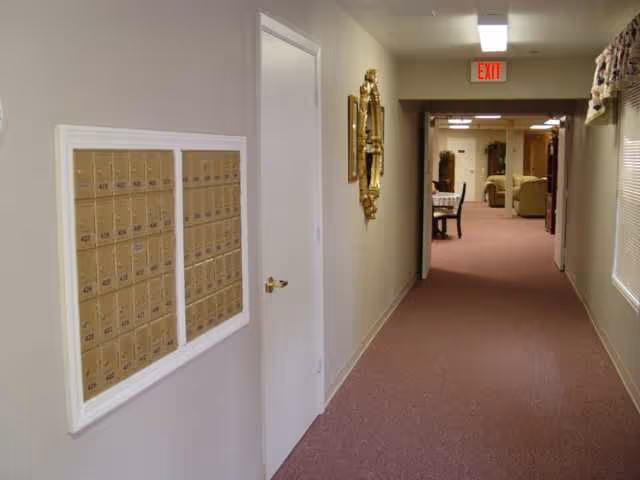 Interior hallway of Southern Wind Assisted Living with beige walls and carpeted floor. On the left wall, there is a set of mailboxes. A white door is next to the mailboxes. Further down the hallway, there is a decorative gold-framed mirror on the wall. At the end of the hallway, there is an exit sign above a doorway leading to a room with chairs and tables.