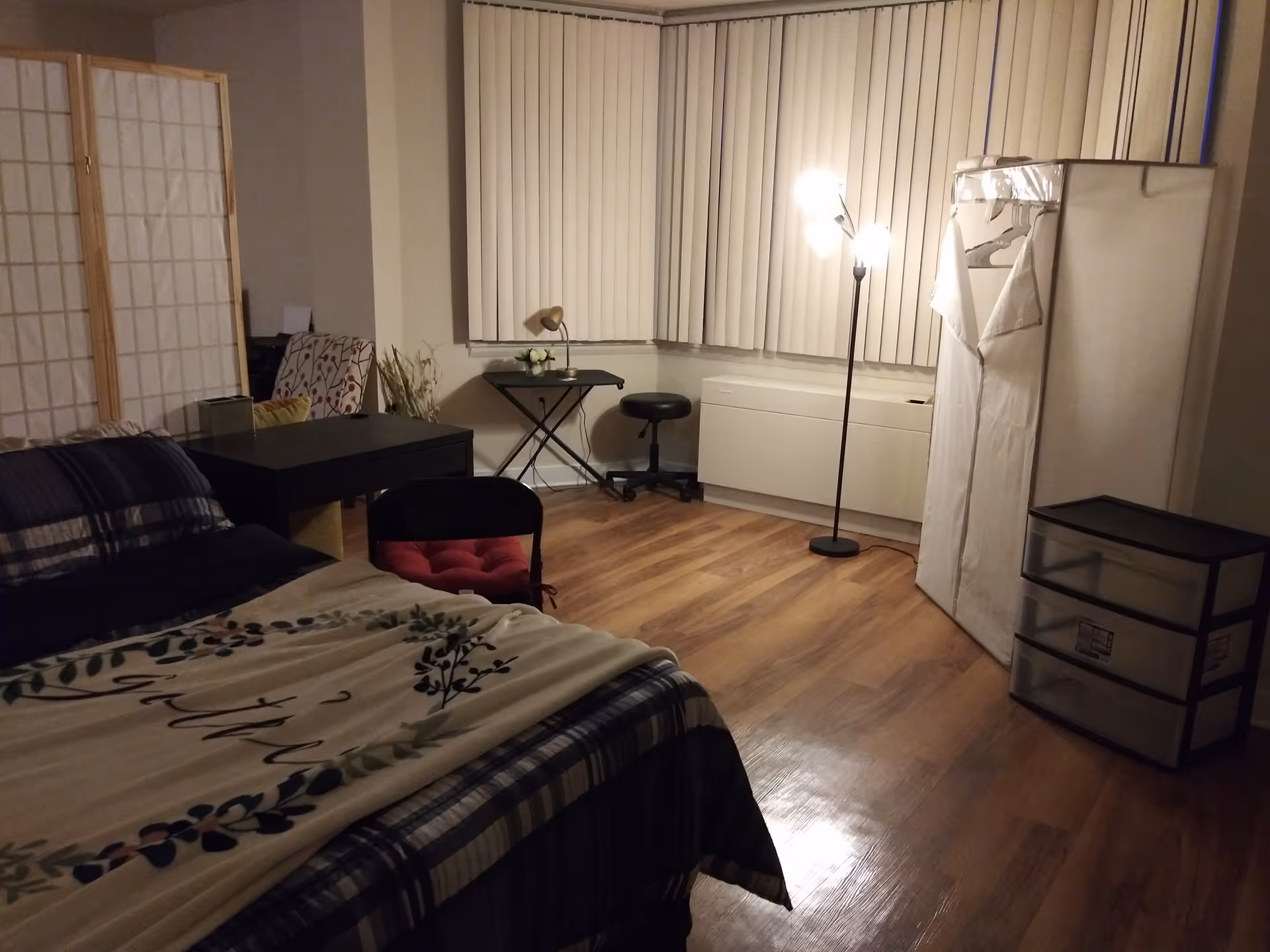 A bedroom with a bed covered in a patterned blanket, a black desk with a chair, a small table with a lamp and flowers, a floor lamp, a fabric wardrobe, and a plastic drawer unit. The room has wooden flooring and vertical blinds covering the windows.