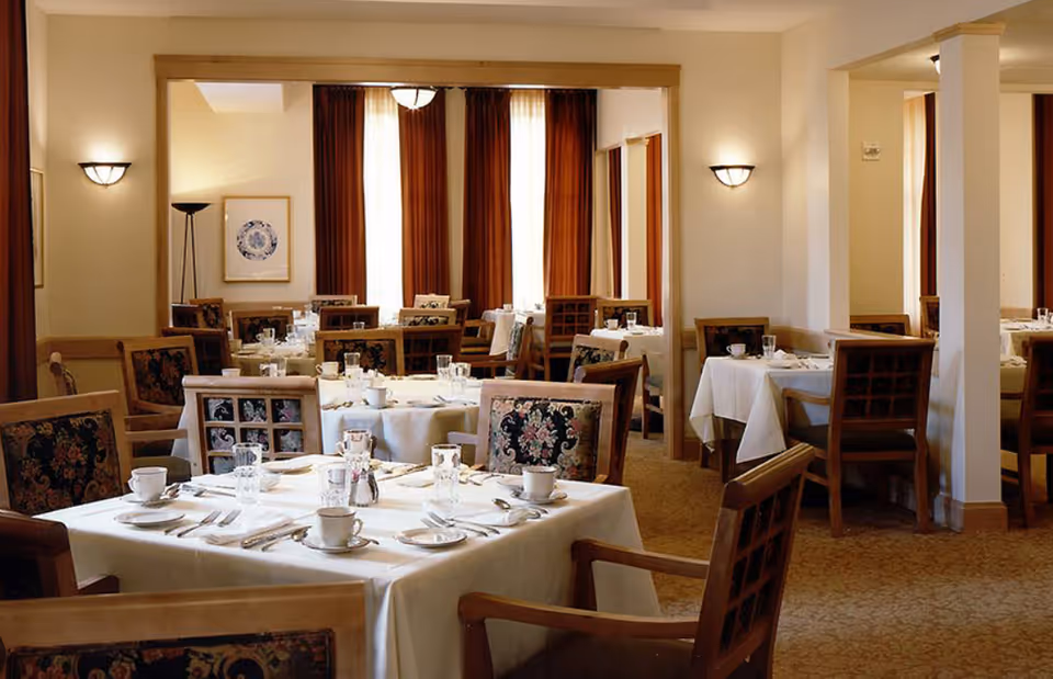 Well-lit dining room with multiple tables set with white tablecloths, cups, silverware, and wooden chairs.