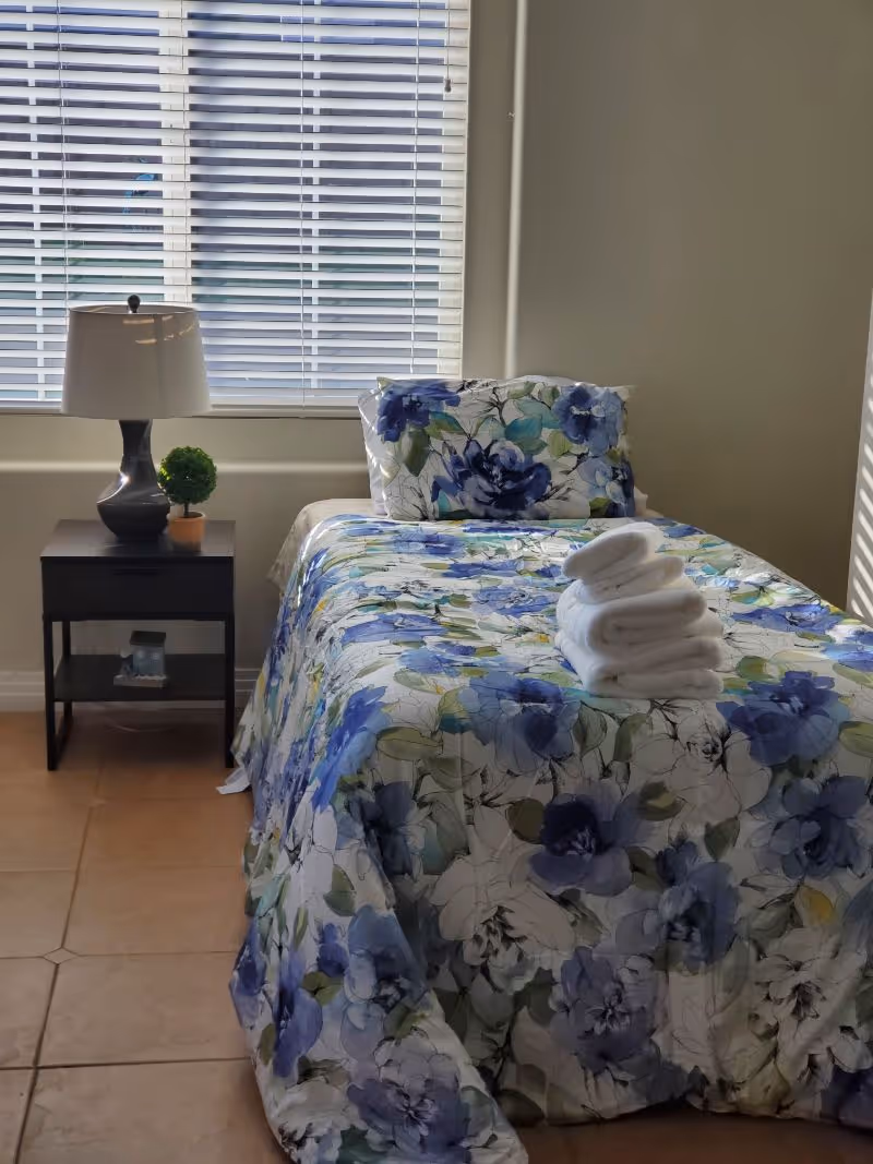 A single bed with a floral bedspread and matching pillow in shades of blue, green, and white. On top of the bed is a neatly folded stack of white towels. Next to the bed is a black nightstand with a small potted plant and a modern table lamp. Behind the bed is a window with white blinds partially closed, allowing some natural light into the room. The floor is tiled in a light brown color.