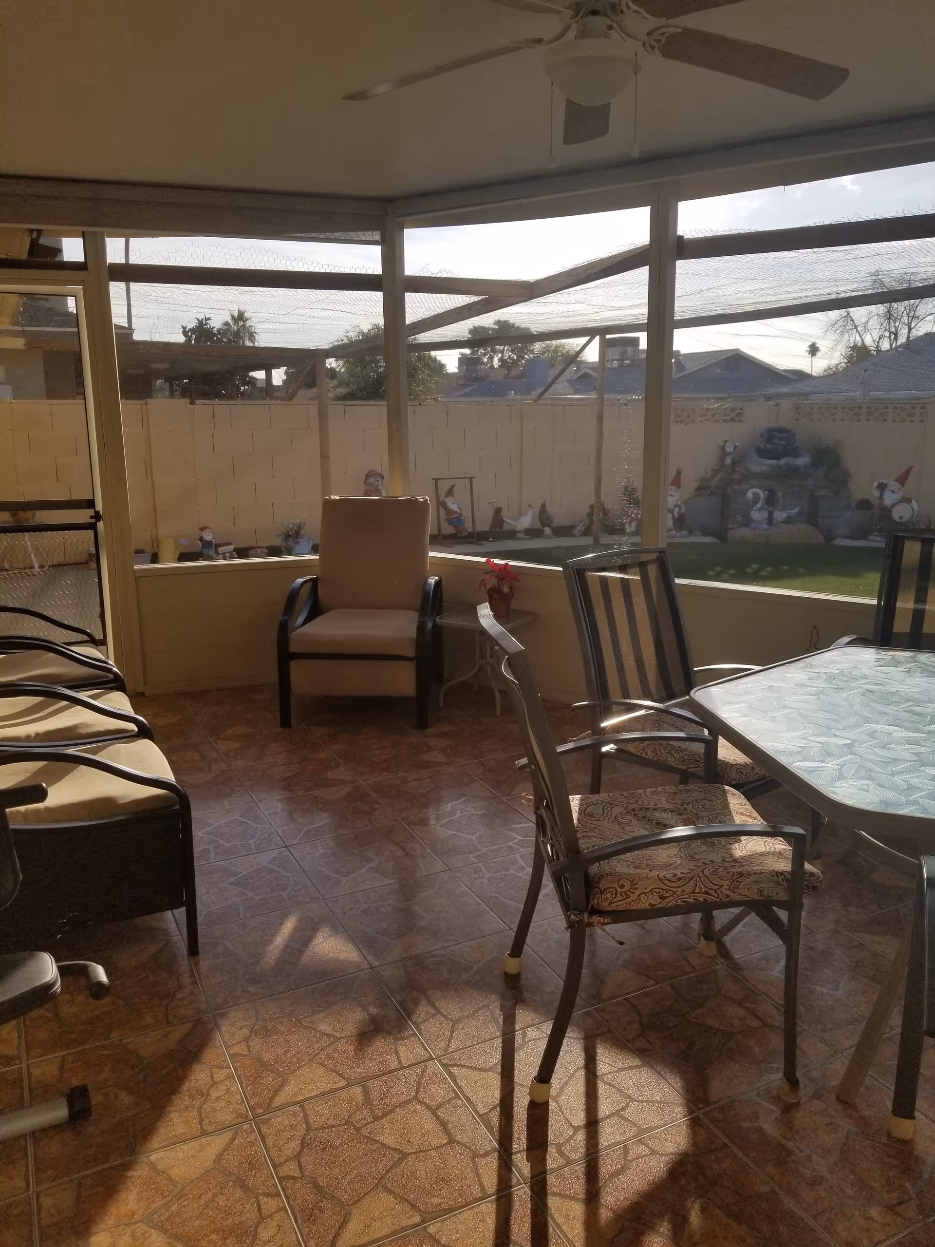 A sunlit enclosed patio area with tiled flooring, featuring a cushioned armchair, a glass-top table with patterned chairs, and a ceiling fan. Outside the screened enclosure, there is a small garden with lawn ornaments including gnomes and a water fountain against a brick wall.