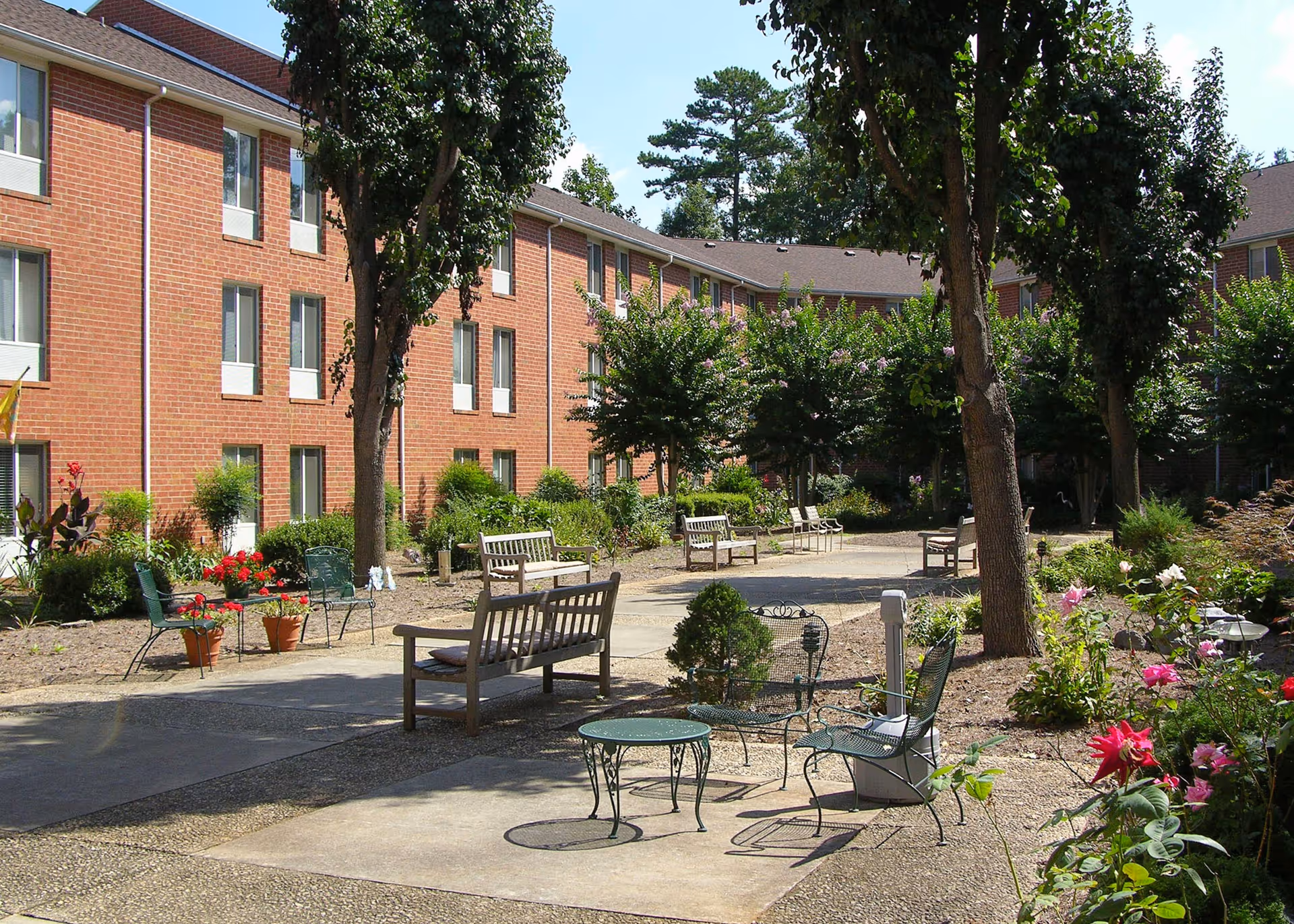 Courtyard with benches, metal tables and chairs and flower beds in front of a three-story brick residential building.
