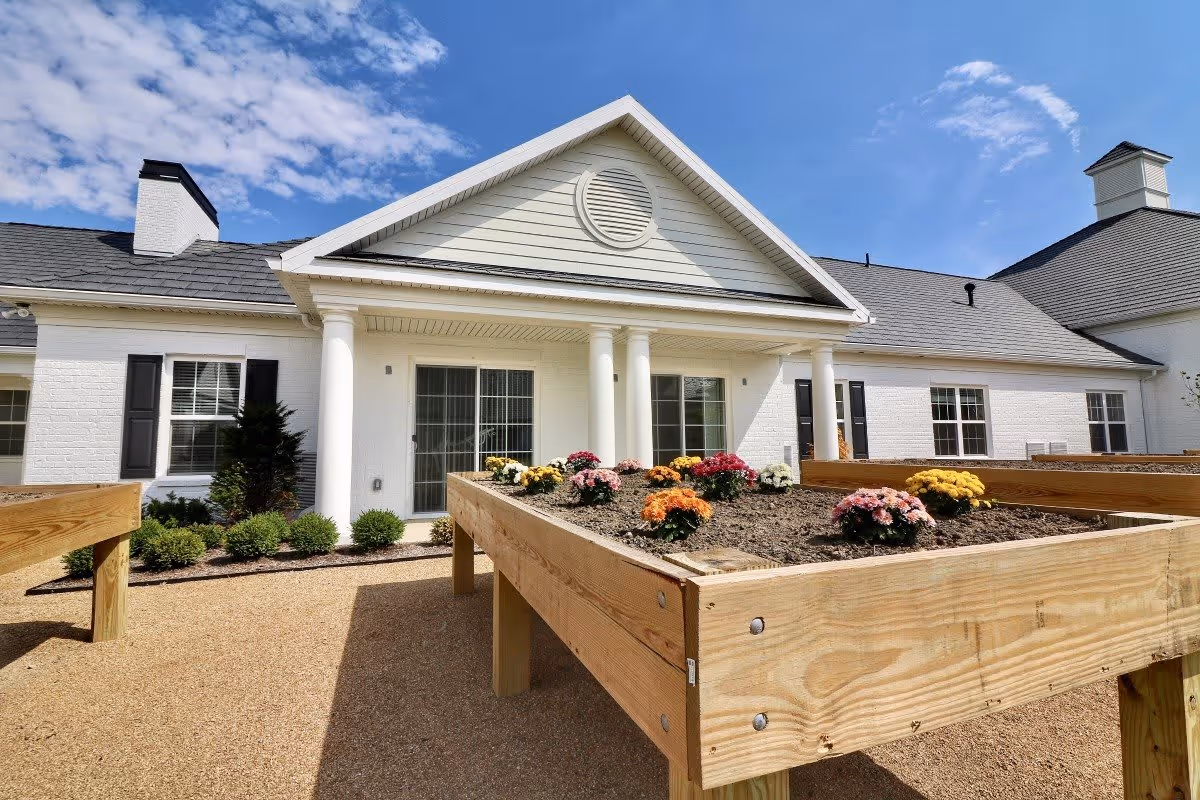Outdoor view of a white senior living facility building with a porch supported by white columns. Raised wooden garden beds with colorful flowers are in the foreground under a partly cloudy blue sky.