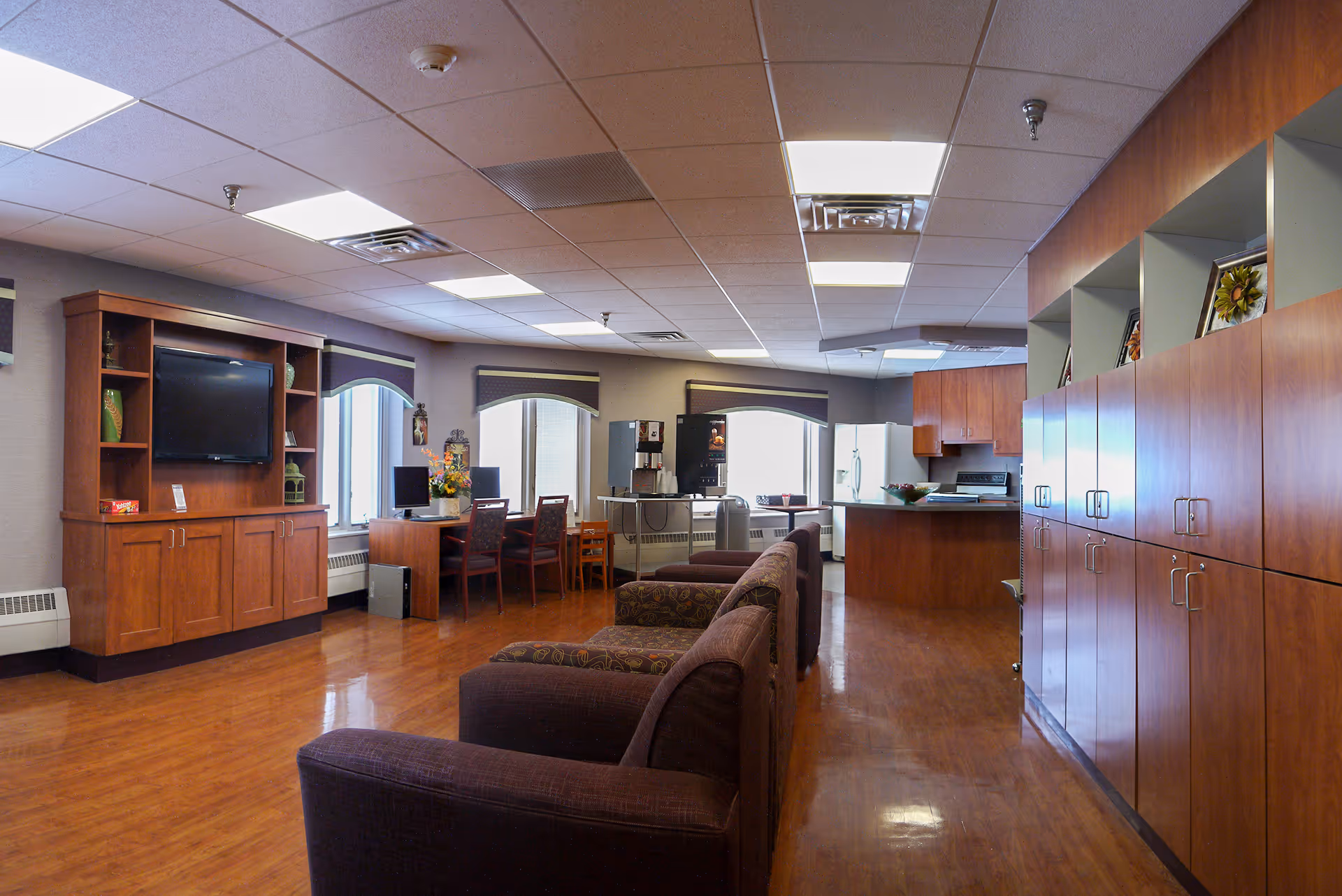 Interior view of a senior living facility common area with wooden flooring, several brown upholstered armchairs, a wooden entertainment center with a TV, a desk with two chairs and computers, a coffee and beverage station, and a kitchen area with wooden cabinets and appliances.