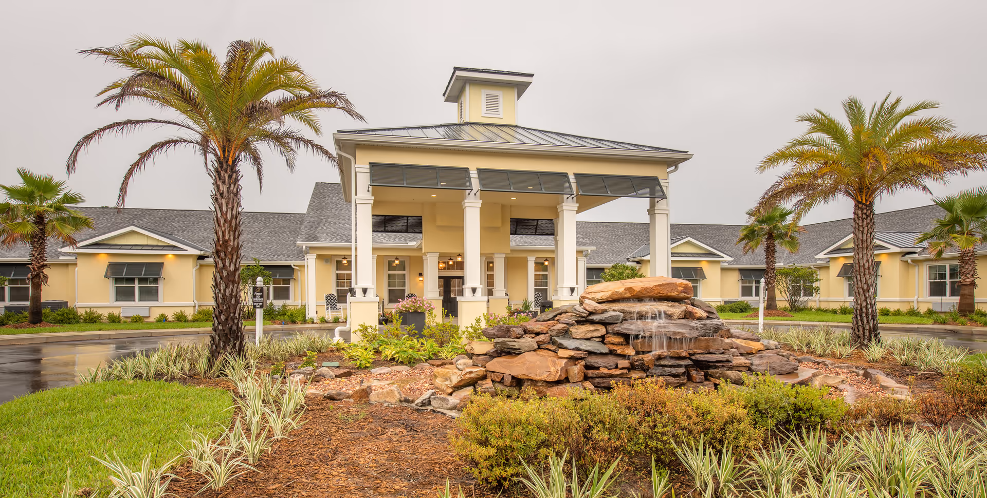 Exterior view of The Addison of Oakleaf senior living facility featuring a covered entrance with white columns, a stone water fountain in the foreground, palm trees, and landscaped greenery under an overcast sky.