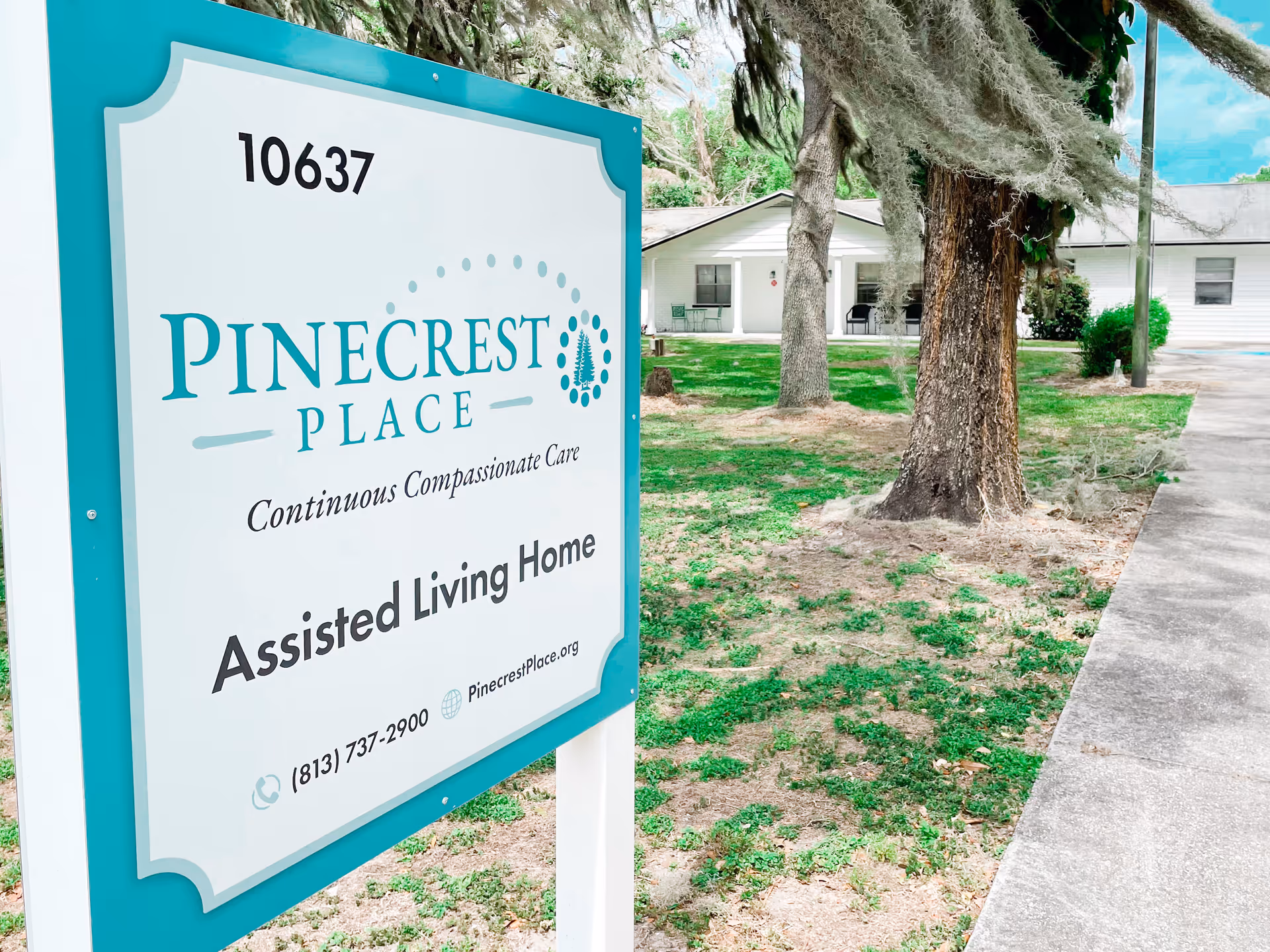 Outdoor view of Pinecrest Place assisted living home sign with the building and trees in the background on a sunny day.