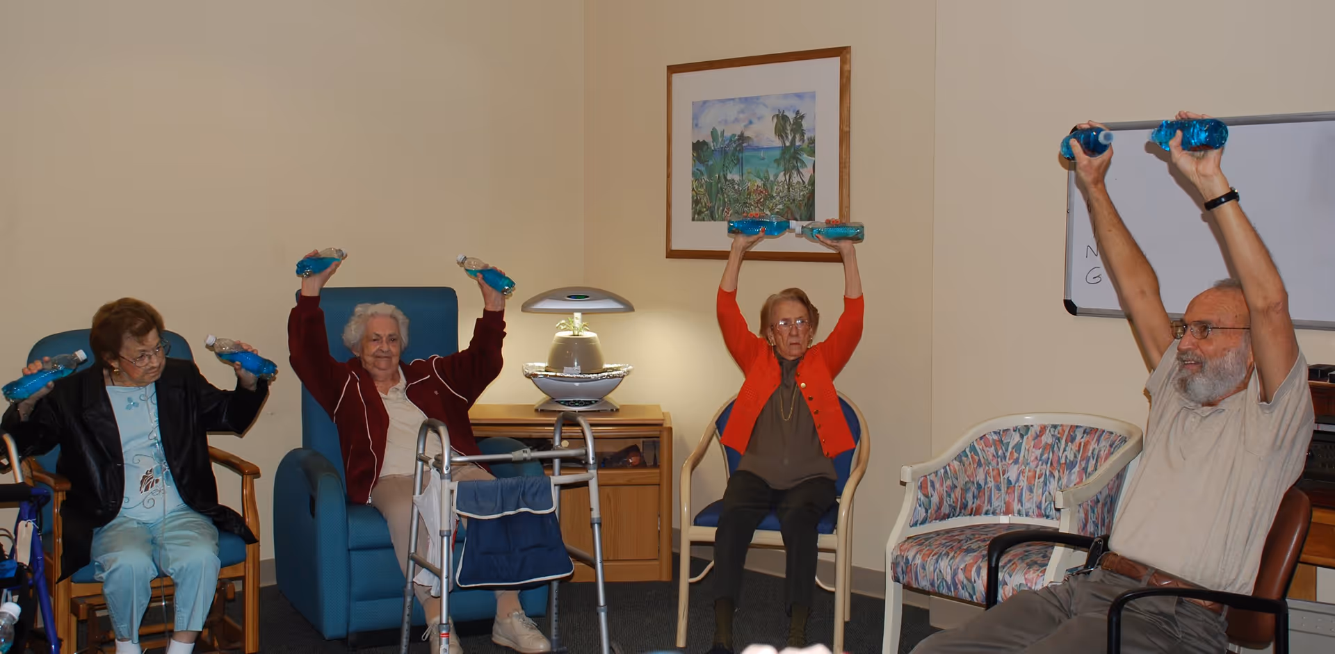 Four elderly individuals seated in chairs in a room, participating in a seated exercise activity by lifting blue water bottles above their heads. The room has beige walls, a framed picture of a tropical scene, a small table with a decorative lamp, and a whiteboard in the background.