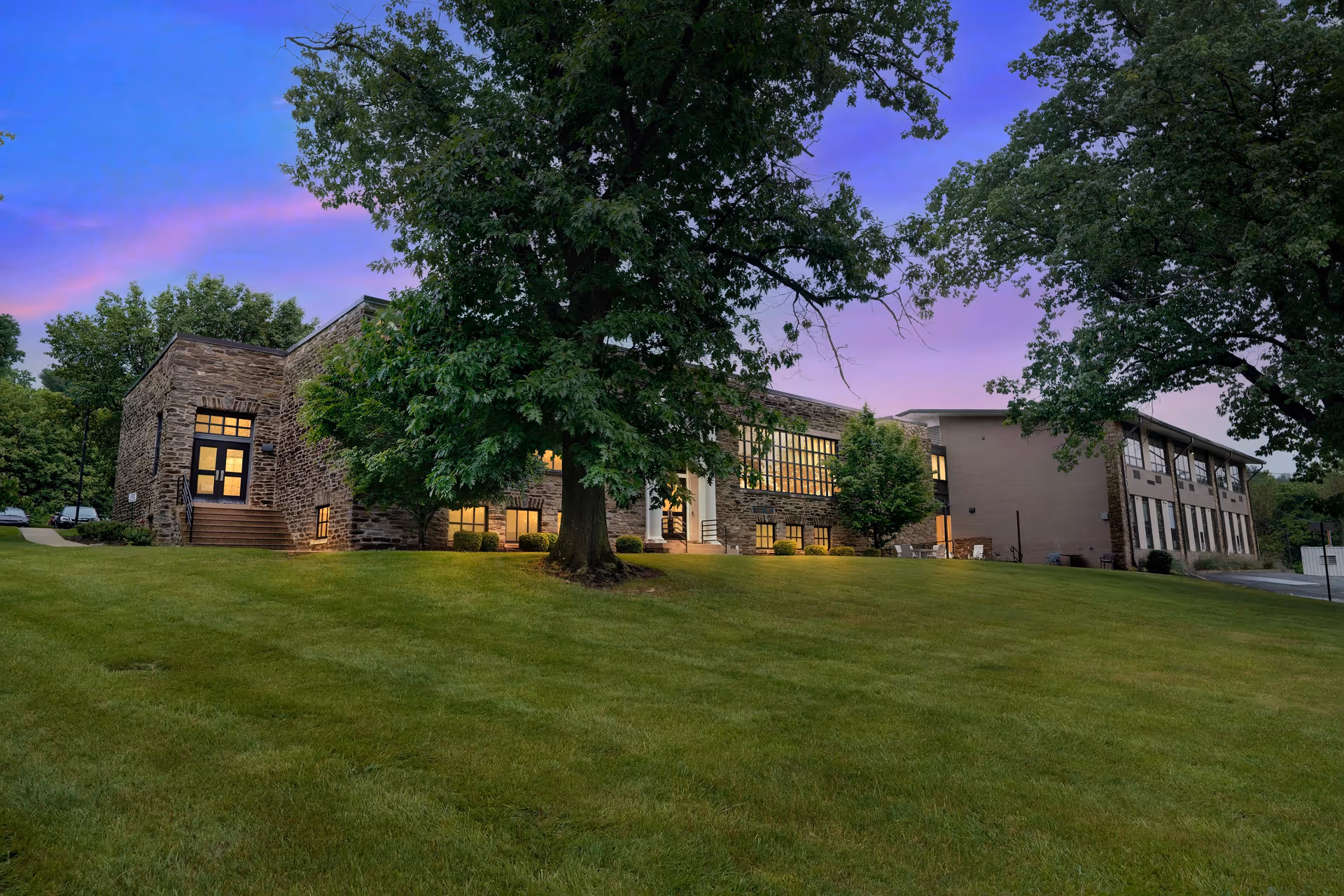 Exterior view of a senior living facility building at dusk with large trees and a well-maintained grassy lawn in the foreground. The building has stone and brick walls with multiple windows illuminated from inside.