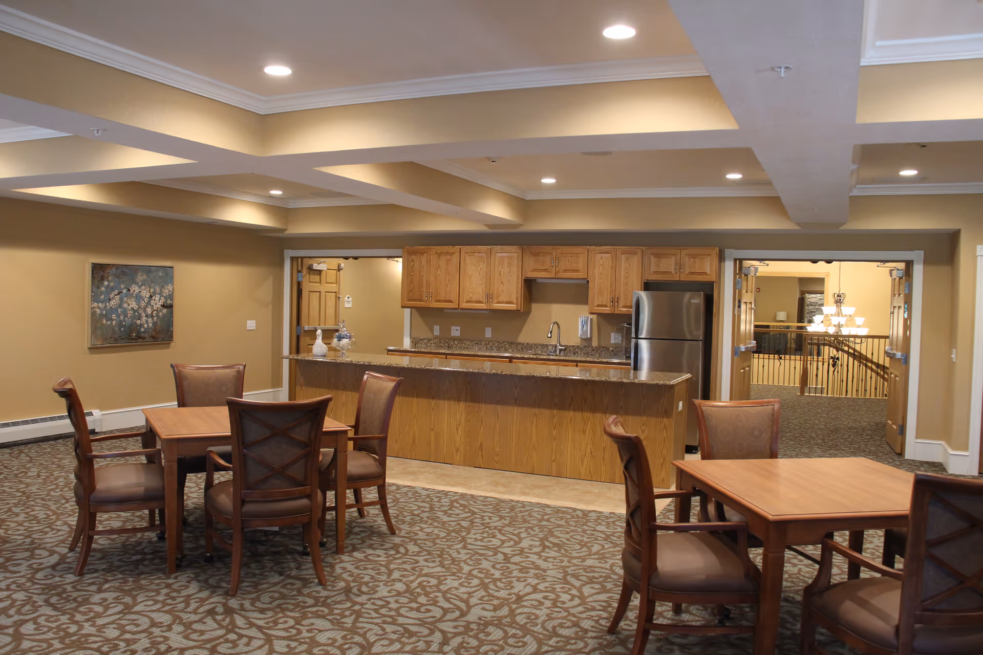 Interior view of a senior living facility common area featuring a kitchen with wooden cabinets, a granite countertop island, and a stainless steel refrigerator. In the foreground, there are two wooden tables each surrounded by four cushioned chairs. The room has beige walls, patterned carpet, recessed lighting, and a painting on the wall to the left.