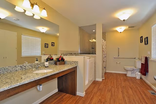 Spacious bathroom with wooden floor, a large granite countertop with a sink and mirror above it, a walk-in shower with glass door, a toilet with grab bars, and a window with blinds. The room is well-lit with ceiling lights and wall-mounted lights above the mirror.