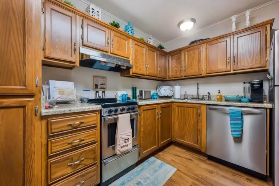 A kitchen with wooden cabinets, a stainless steel stove with a towel hanging on the handle, a dishwasher with a blue towel, a refrigerator, and various kitchen items on the countertops including a clock, paper towel holder, and coffee maker.