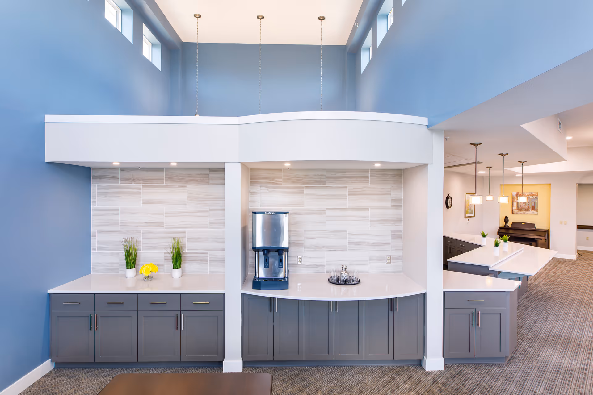 Bright common area kitchenette with gray cabinets, a coffee machine, potted plants, and blue walls under a high ceiling.