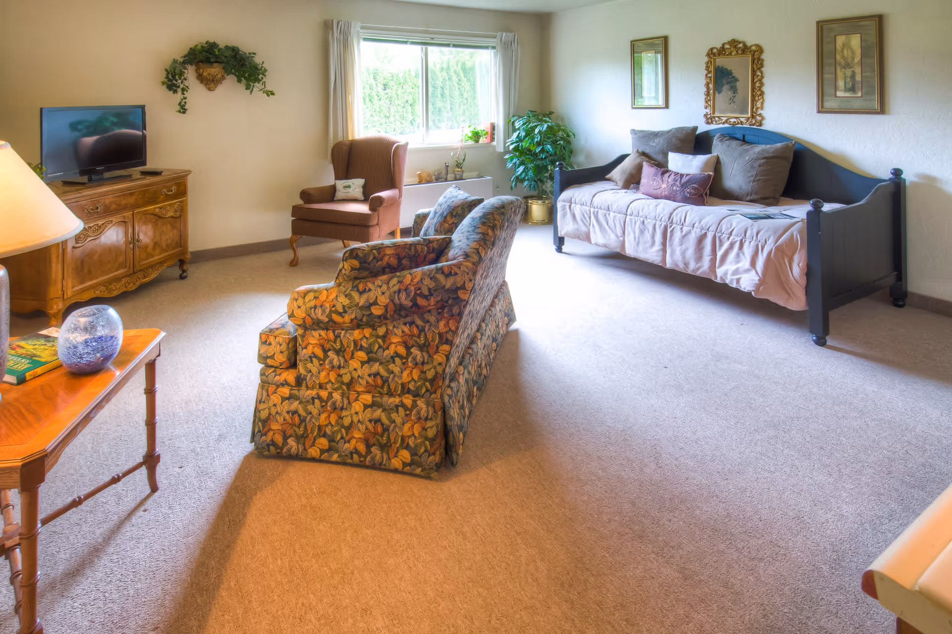 A cozy living room with a floral patterned armchair, a brown wingback chair, a daybed with multiple pillows, a wooden TV stand with a flat screen TV, a wooden side table with a decorative glass orb and books, a window with white curtains, and framed artwork on the walls.