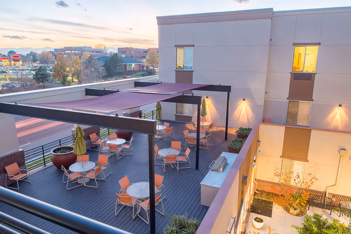 Outdoor patio area at Village at Belmar with several round tables and orange chairs under purple shade sails, large potted plants, and a view of nearby buildings and parking lot at sunset.