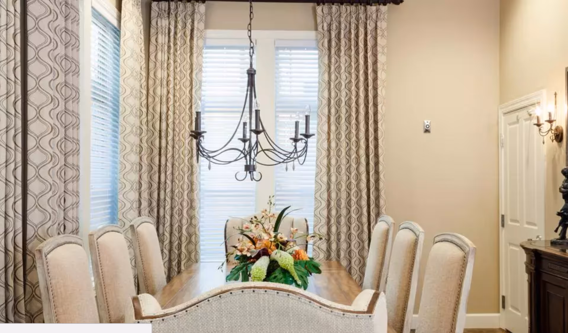 Dining room with a wooden table, upholstered chairs, a chandelier, patterned curtains, and a floral centerpiece.