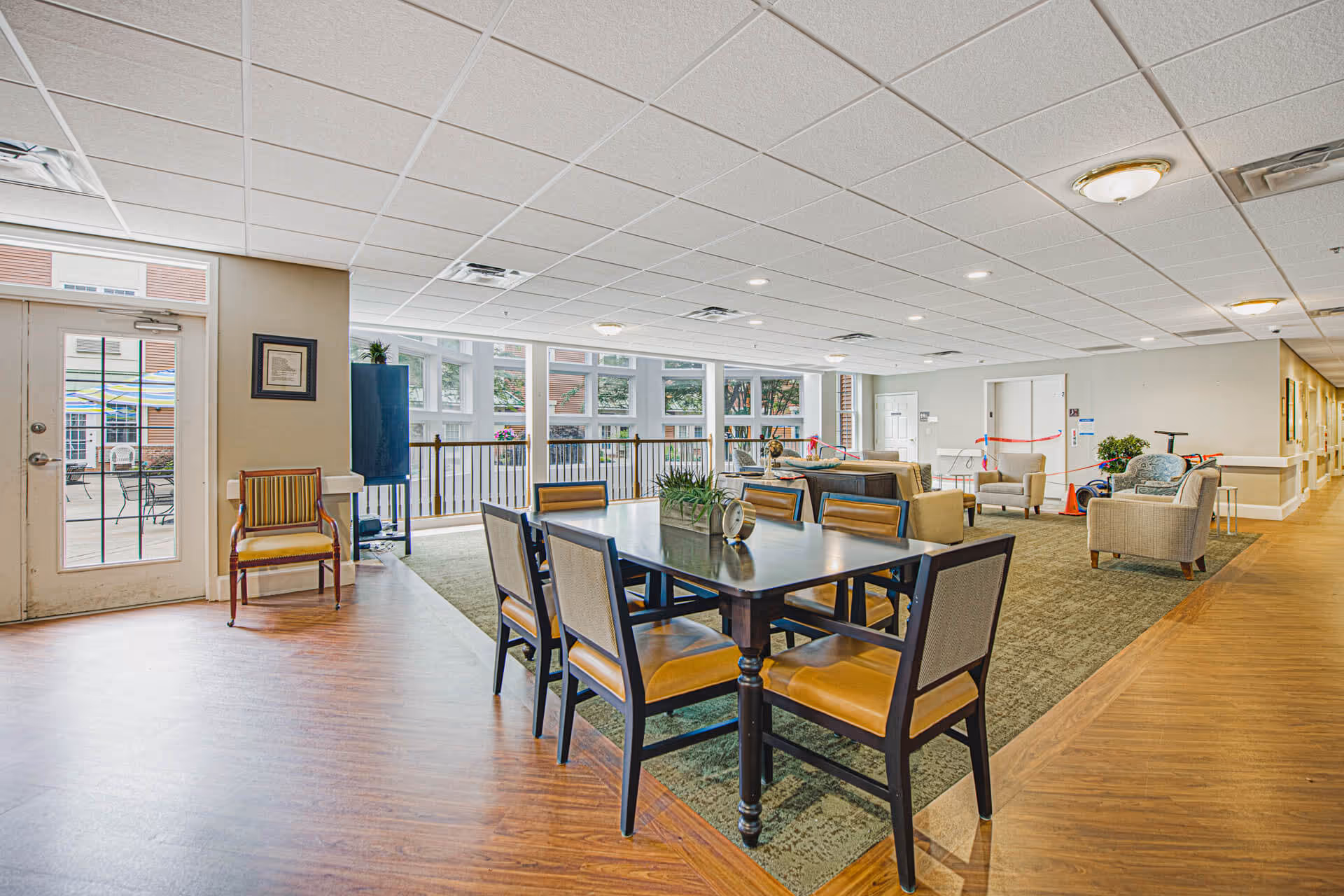 A spacious and well-lit common area in a senior living facility with a rectangular dining table surrounded by six chairs in the foreground. The room features large windows letting in natural light, a seating area with sofas and armchairs, and a door leading to an outdoor patio with tables and umbrellas. The floor is a combination of wood and carpet, and the ceiling has recessed lighting.