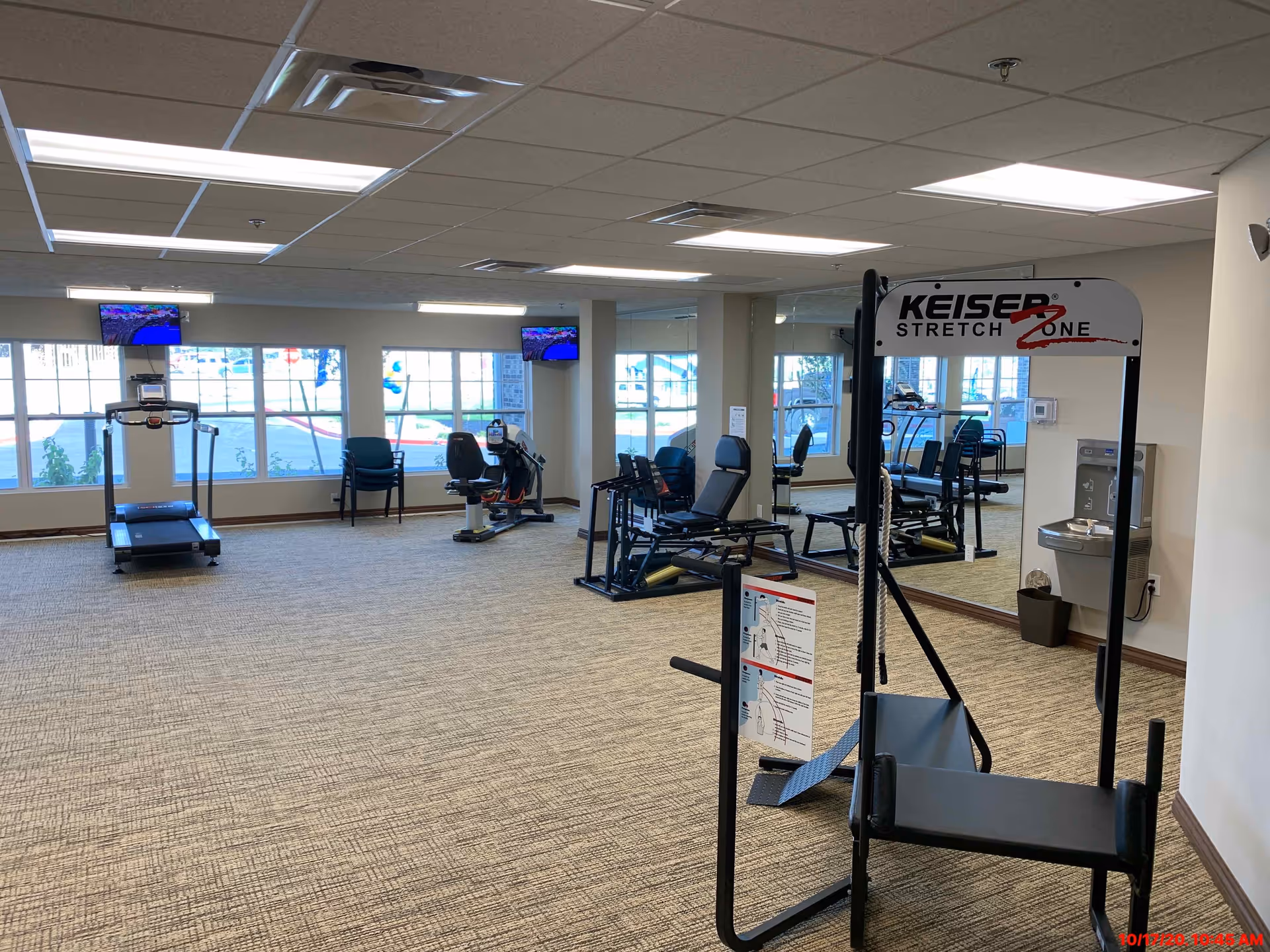 A bright interior fitness room with treadmills, recumbent bikes, strength machines, mirrors, and a Keiser Stretch Zone unit.