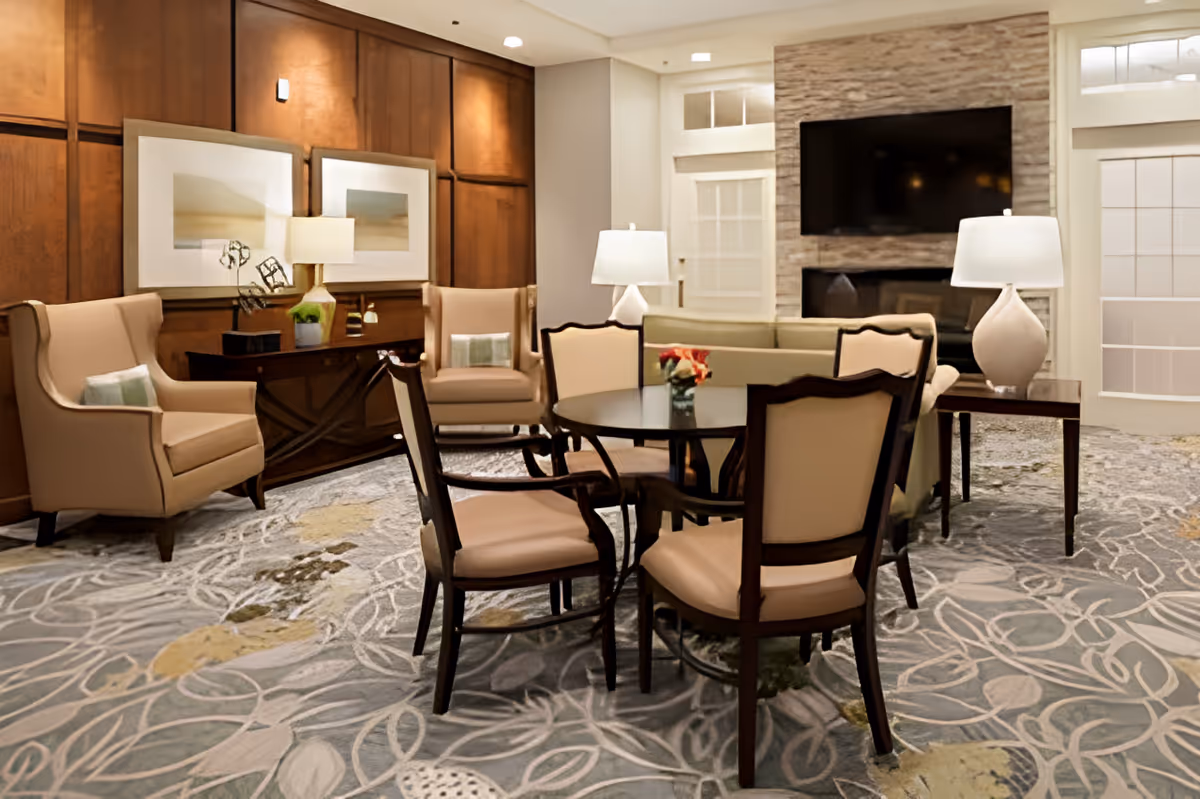 Well-appointed communal lounge with upholstered chairs around a round table, lamps, and a wall-mounted TV above a fireplace.