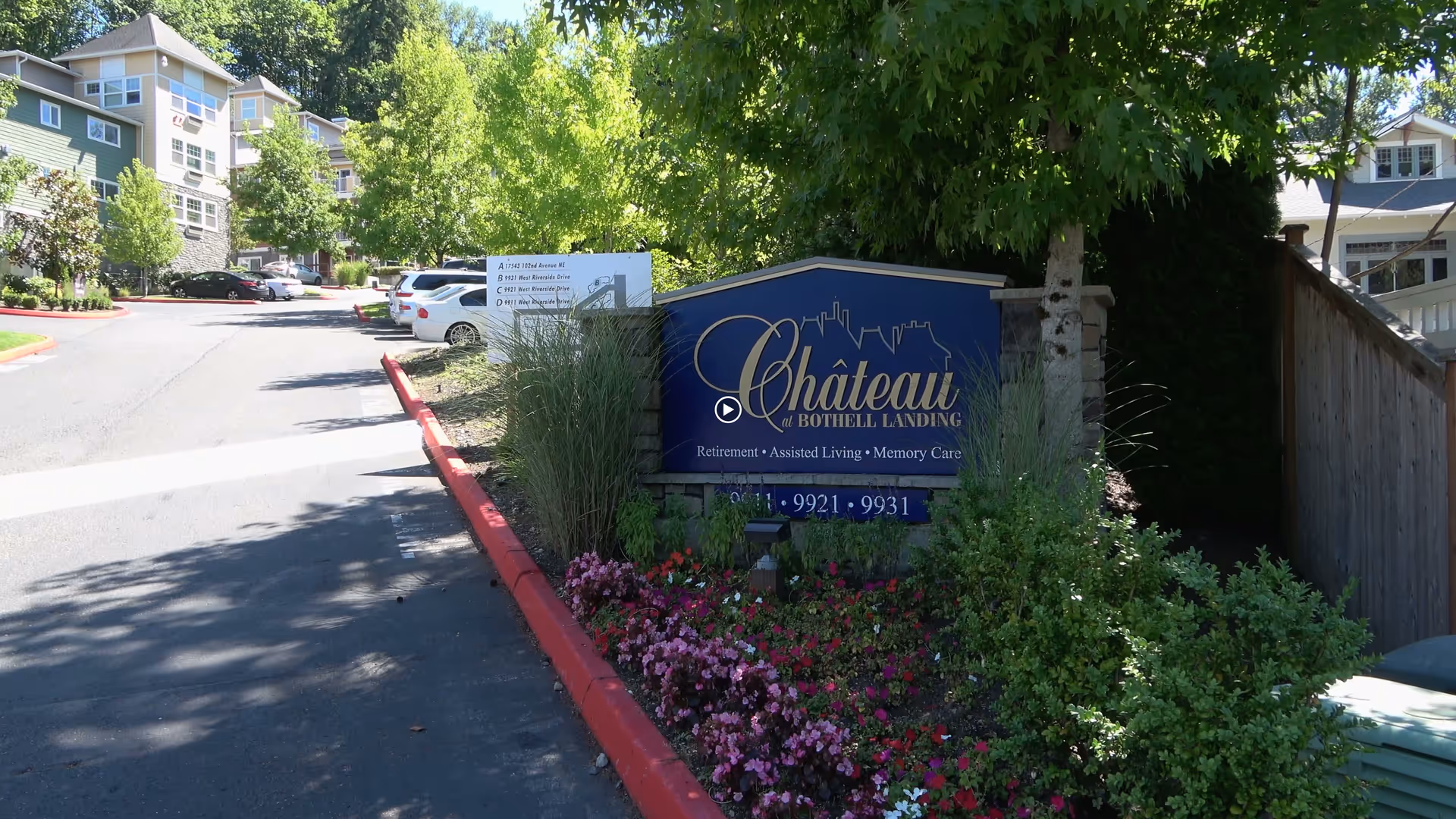 Entrance sign for Chateau at Bothell Landing Retirement community surrounded by flowers and greenery, with a driveway and residential buildings visible in the background.