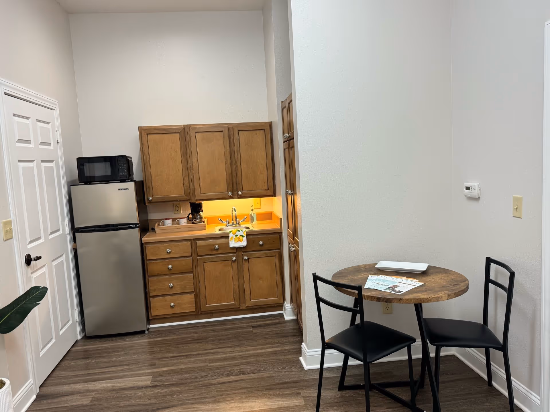 A small kitchen area with wooden cabinets, a stainless steel refrigerator with a microwave on top, a sink with a coffee maker nearby, and a round wooden table with two black chairs. The floor is wood, and the walls are white.