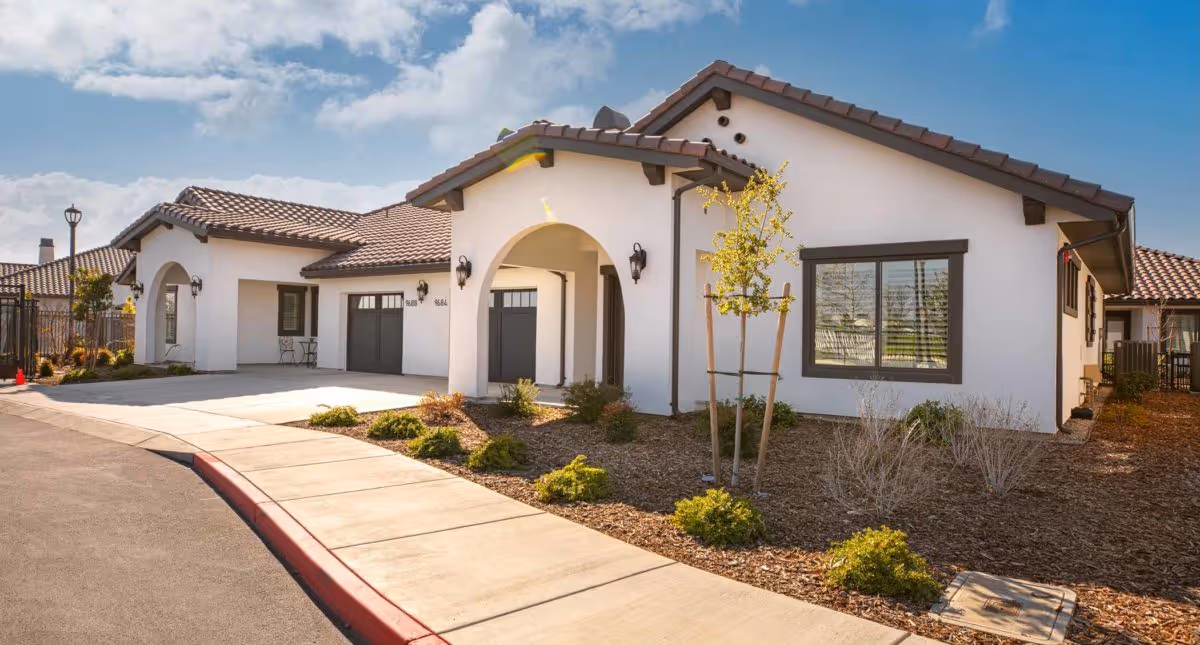 Single-story Mediterranean-style building with white stucco walls, tiled roof, garage doors and landscaped walkway under a partly cloudy sky.