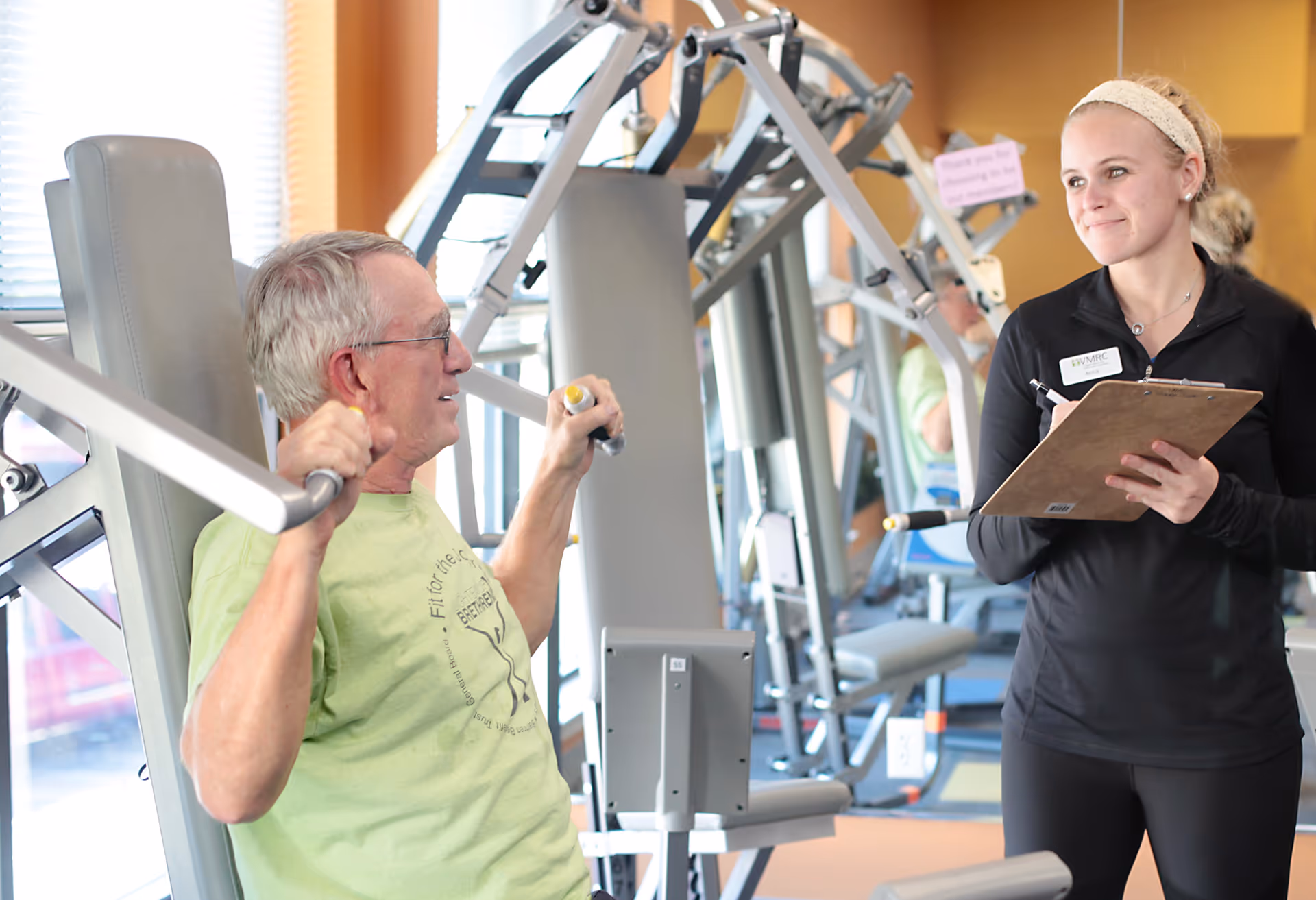 An elderly man in a green t-shirt is using a chest press machine in a gym while a female fitness trainer with a clipboard observes and smiles. The gym has exercise equipment and large windows letting in natural light.