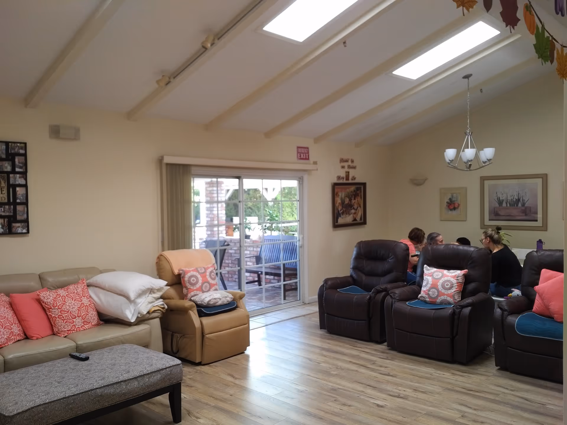 A cozy living room area in an assisted living facility with beige and brown recliner chairs, a beige sofa with red and patterned pillows, a cushioned ottoman, and a group of people sitting and talking near the back wall. The room has wooden flooring, a sliding glass door leading outside, and framed artwork on the walls. A chandelier hangs from the ceiling, and there are autumn leaf decorations hanging near the ceiling.