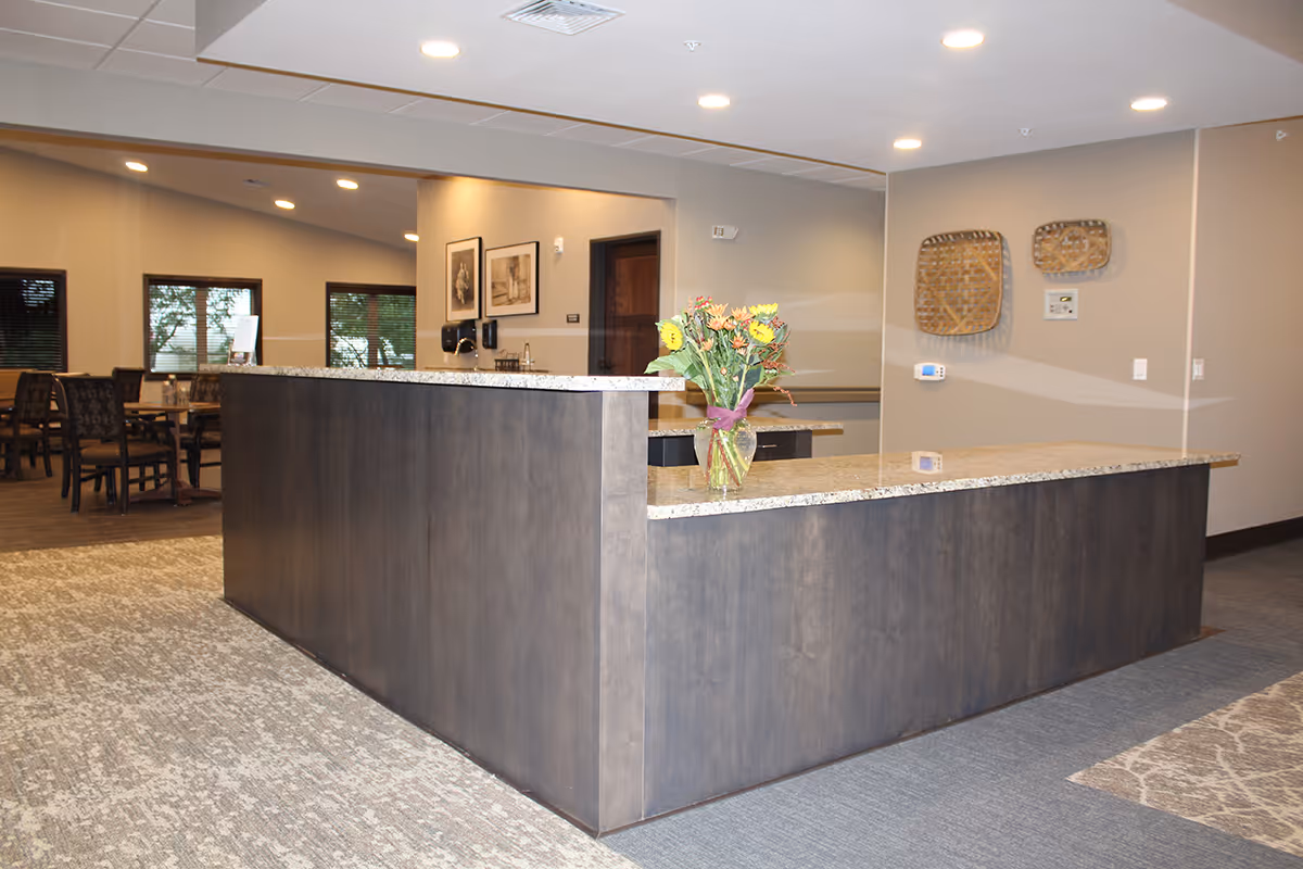 Reception desk area with a granite countertop and a vase of flowers on it, in a senior living facility. In the background, there are dining tables and chairs near windows with blinds. The walls are decorated with framed pictures and woven baskets.