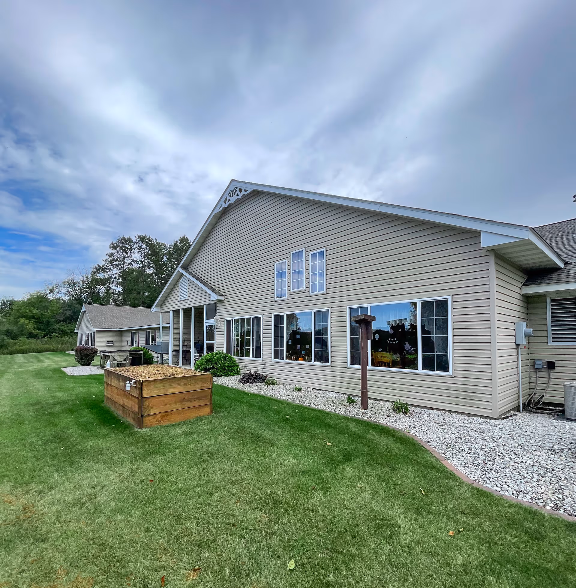 Exterior view of a beige siding building with multiple windows and a sloped roof under a cloudy sky. The building is surrounded by a well-maintained green lawn with a wooden raised garden bed and a bird feeder in the foreground. Trees and shrubs are visible in the background.