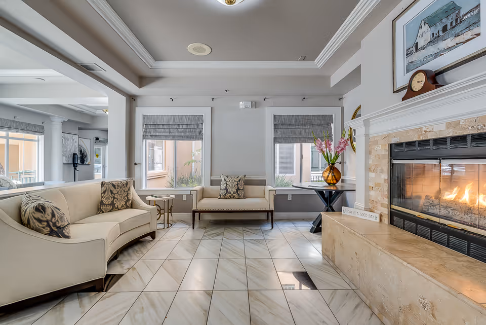 A bright and elegant living room area with a curved beige sofa and a matching loveseat, both adorned with patterned throw pillows. The room features large windows with gray Roman shades, a fireplace with a lit fire, a mantel clock, and a framed painting above it. A round table with a vase of pink flowers is placed near the fireplace. The floor is tiled with a light marble pattern, and the ceiling has a tray design with crown molding.
