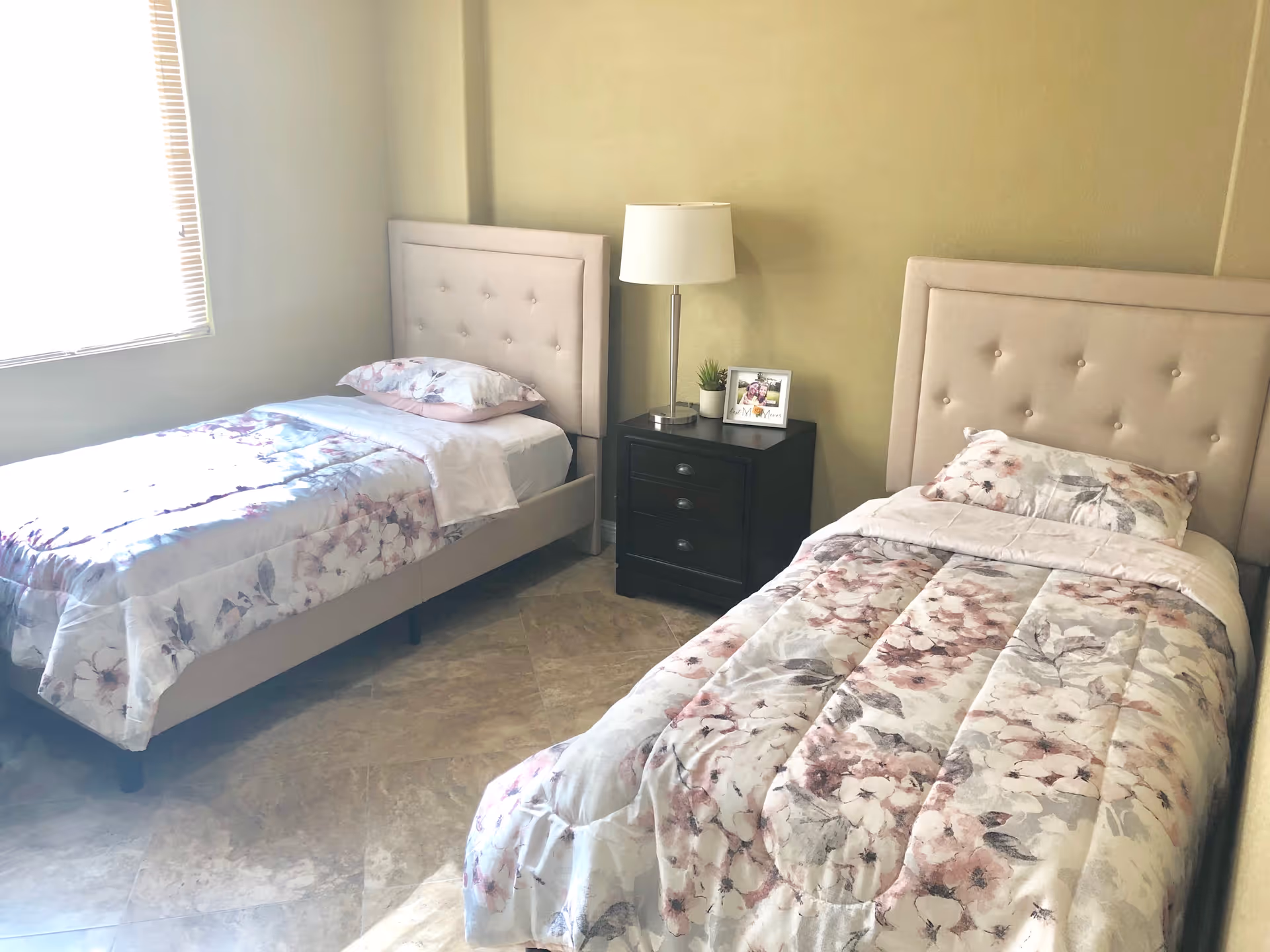 A bedroom with two single beds featuring beige tufted headboards and floral-patterned bedding. Between the beds is a black nightstand with three drawers, a white table lamp, a small potted plant, and a framed photo. A window with blinds allows natural light into the room.
