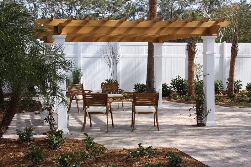 Outdoor seating area with a wooden pergola supported by white columns. Four wooden chairs with white cushions are arranged around a small round table on a paved surface. The area is surrounded by plants, small trees, and a white fence in the background.