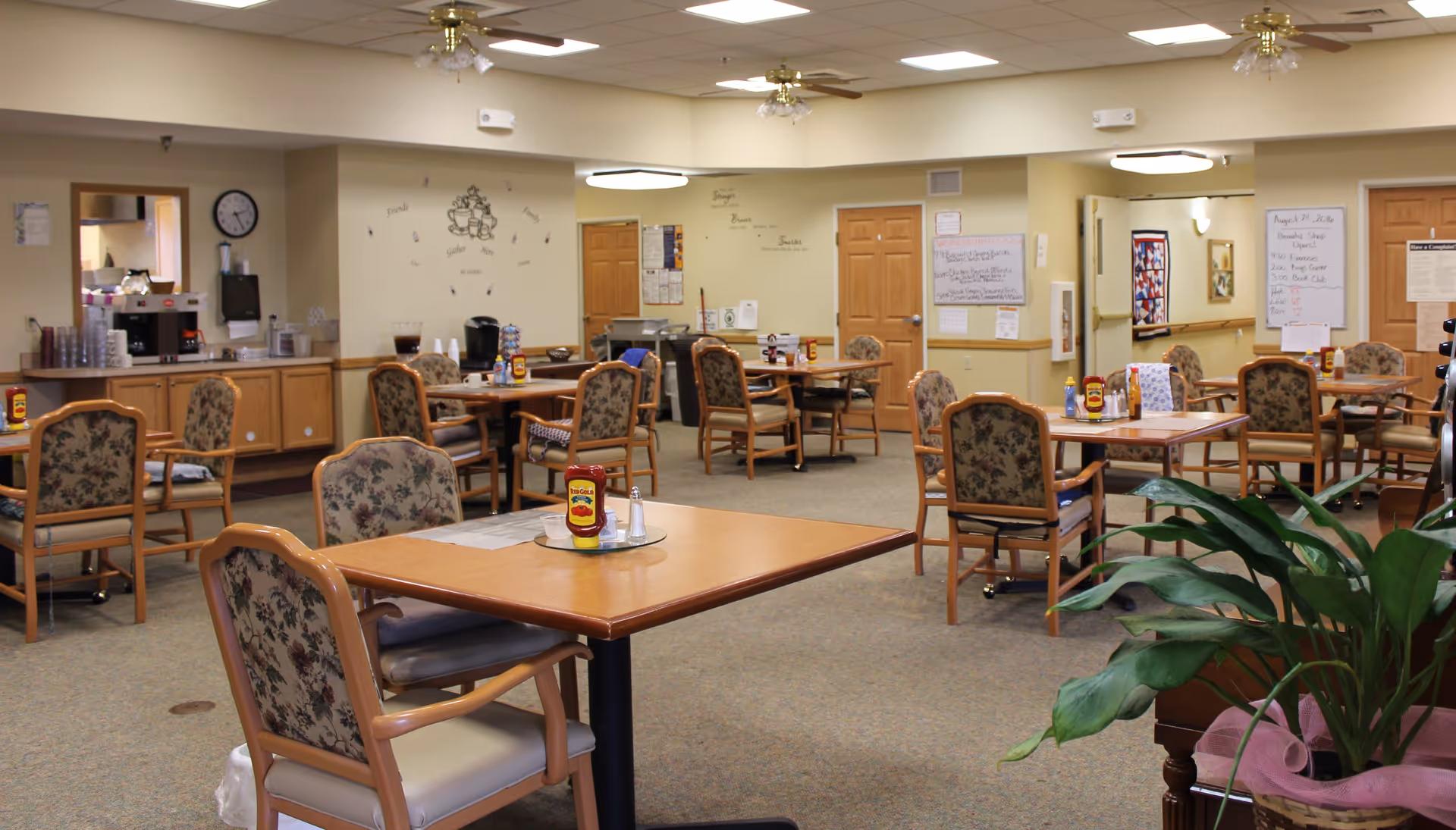 A spacious dining room in an assisted living facility with several wooden tables and floral upholstered chairs. Each table has condiments like ketchup and salt shakers. The room has beige walls, ceiling fans with lights, and a carpeted floor. There is a counter area with cups and a coffee machine on the left side, and various notices and decorations on the walls.
