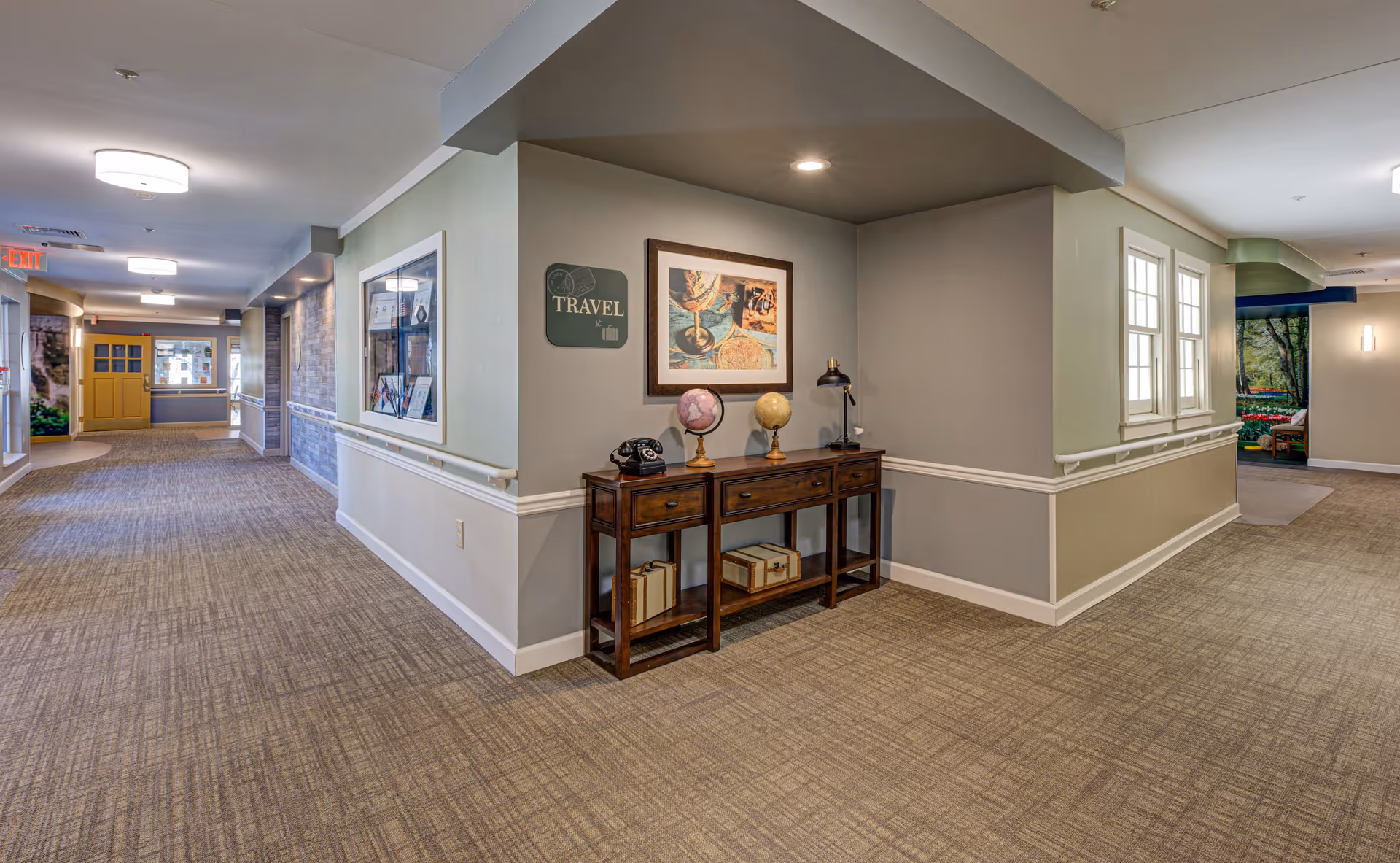 Interior hallway of Arden Courts - ProMedica Memory Care Community (Whippany) with beige carpet, light gray walls, and white trim. A wooden console table against the wall holds two globes, a vintage telephone, a lamp, and some decorative boxes underneath. A framed travel-themed picture hangs above the table, and a sign labeled 'TRAVEL' is mounted on the wall. The hallway extends with doors and windows visible, and there is a yellow door at the far end.