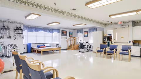 Bright rehabilitation therapy room with exercise equipment, treatment tables, chairs and mobility aids.