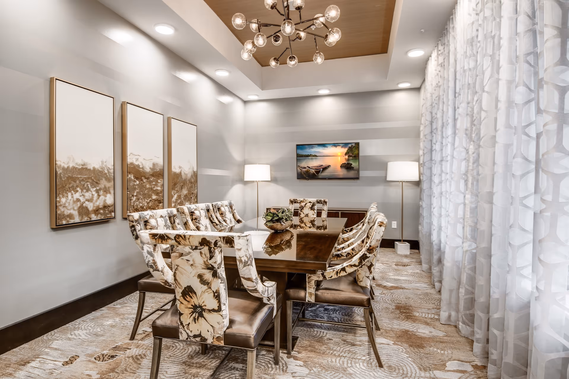 A modern dining room with a rectangular dark wood table surrounded by eight floral upholstered chairs. The room features a patterned carpet, three abstract paintings on the left wall, two floor lamps, a flat-screen TV mounted on the far wall displaying a sunset over a lake, and large sheer curtains on the right side. A contemporary chandelier with multiple round bulbs hangs from the ceiling.