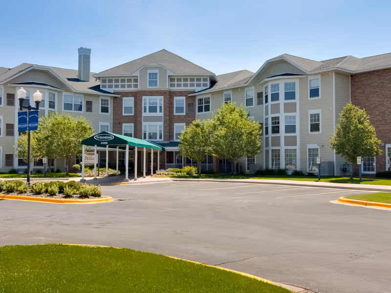 Exterior view of a multi-story senior living facility named Waterford with a covered entrance, surrounded by trees and a parking lot under a clear blue sky.