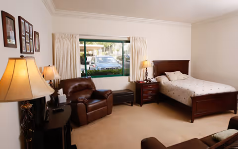 A furnished bedroom with a single bed, nightstands and lamps, a leather recliner, and a window looking out toward parked cars.