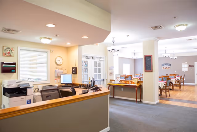 Reception area with a desk, computer, printer, and office supplies in the foreground. In the background, a dining area with tables and chairs is visible, along with windows and light fixtures on the ceiling.
