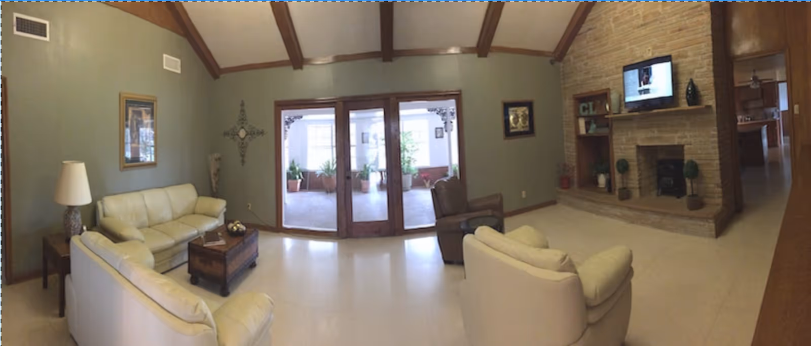 A cozy living room with cream-colored sofas and a brown armchair arranged around a wooden coffee table. The room features a brick fireplace with a flat-screen TV mounted above it, wooden beams on the ceiling, and large glass doors leading to a sunlit indoor garden area with potted plants.