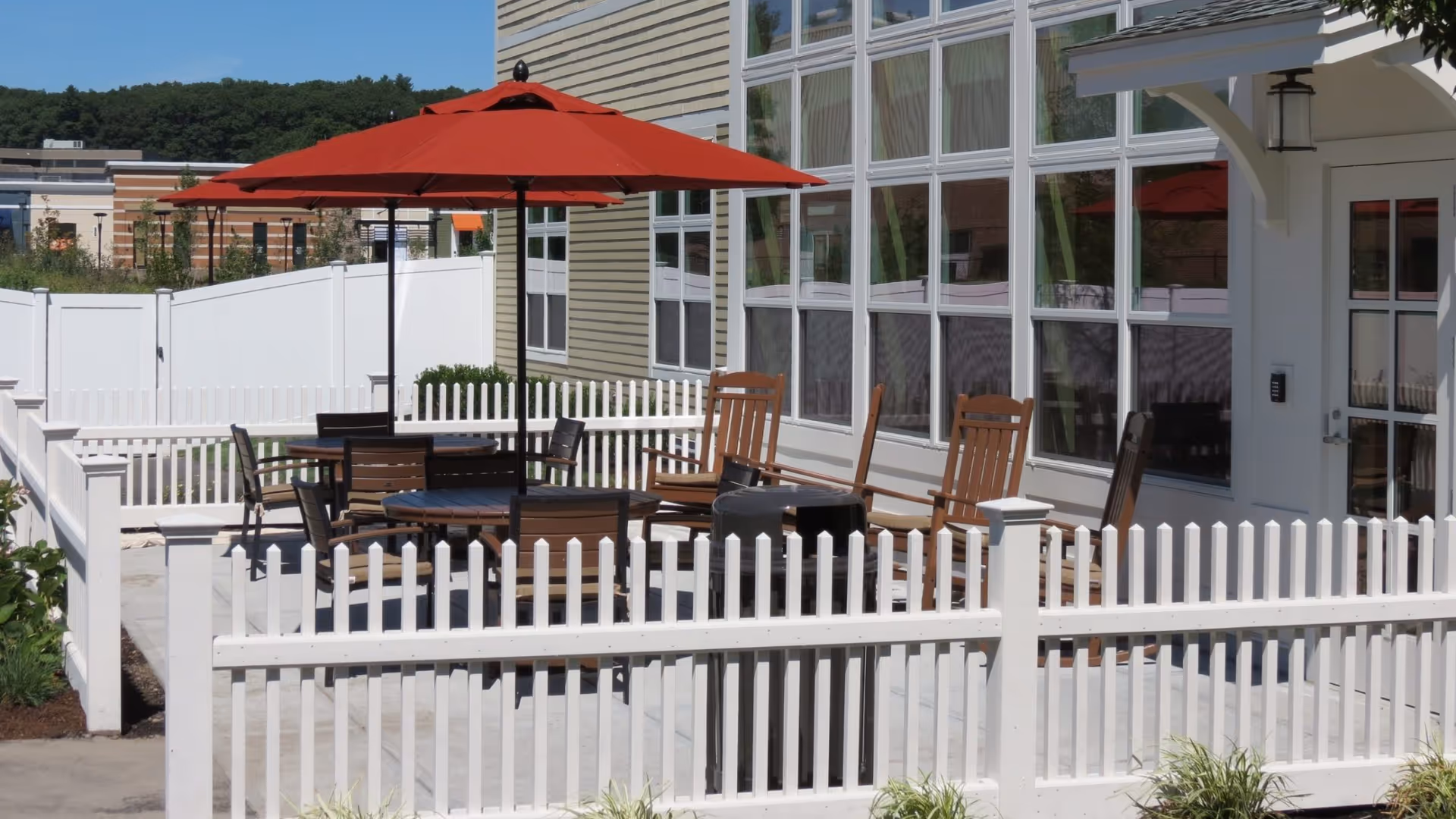 Outdoor patio area with wooden chairs and tables under red umbrellas, surrounded by a white picket fence, next to a building with large windows and a door.