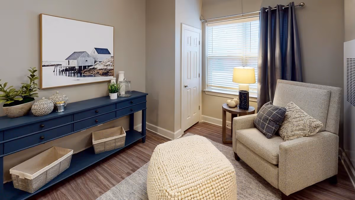 A cozy living room corner featuring a beige armchair with two decorative pillows, a round wooden side table with a lamp and small pumpkins, a large knitted ottoman, a blue console table with plants and decorative items, and a framed picture of houses on stilts by the water hanging on the wall.
