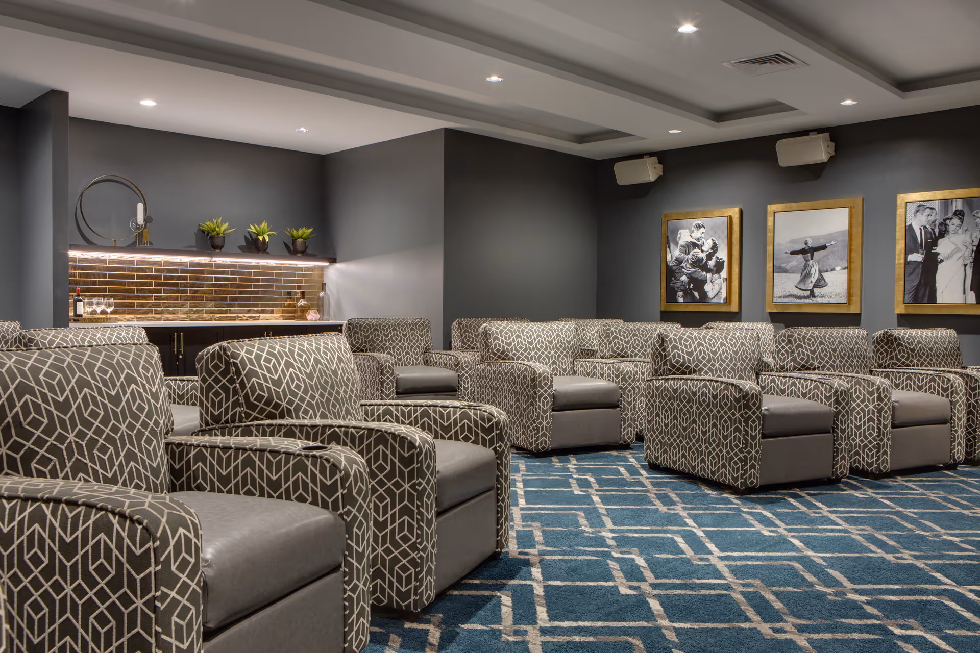 A cozy lounge area with multiple patterned armchairs arranged in rows on a blue carpet with a geometric design. The walls are painted gray and decorated with three framed black and white photographs. There is a small bar area with a brick backsplash, a round mirror, and several small potted plants on a shelf.