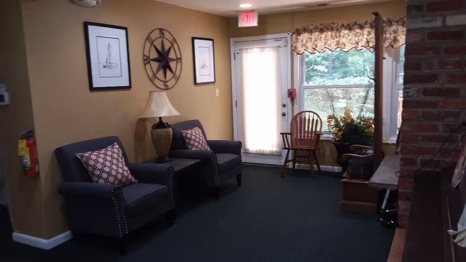 A cozy interior seating area with two dark armchairs featuring patterned cushions, a side table with a lamp between them, framed nautical-themed artwork and a decorative compass on the wall. There is a door with a sheer curtain, a wooden chair near a window with floral valance, and a brick fireplace partially visible on the right.