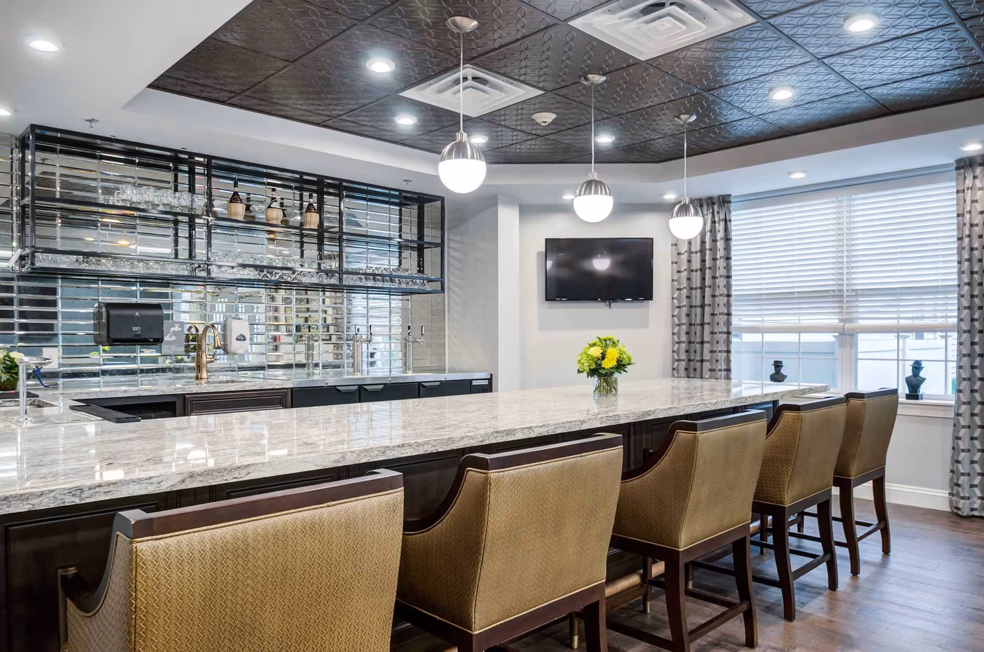 Modern dining area with a long marble countertop bar, six upholstered bar stools, pendant lights hanging from a decorative ceiling, a wall-mounted TV, and a large window with patterned curtains. Behind the bar is a mirrored backsplash with shelves holding glassware and decorative items.