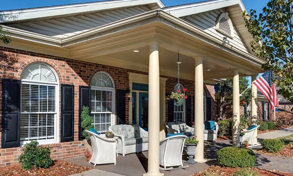 Front entrance of a brick senior living building with a covered porch, columns, outdoor seating, and an American flag.