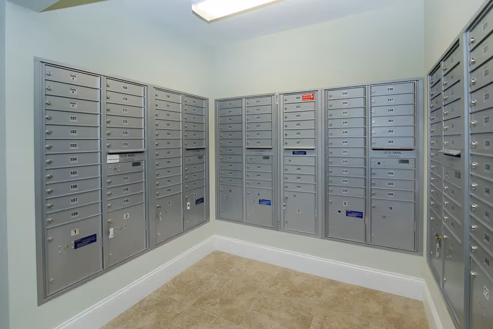 A small room with multiple gray metal mailboxes mounted on two adjacent walls, each labeled with numbers and some with keys inserted. The floor is tiled with beige tiles and the walls are painted light gray. The room is well-lit with a ceiling light.