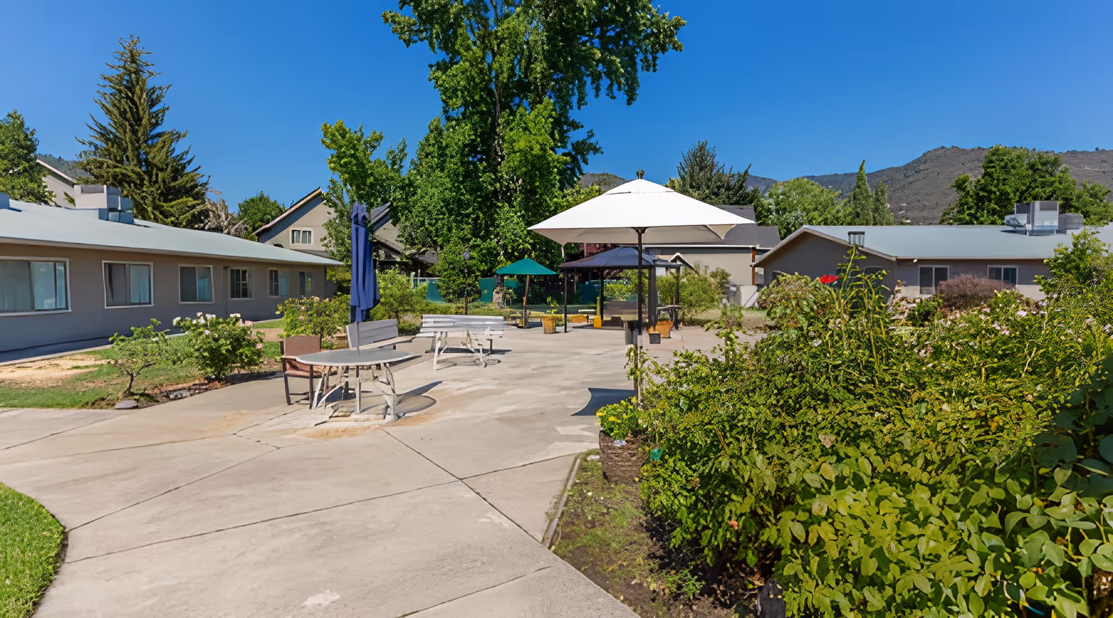 Outdoor patio area at a senior living facility with concrete walkways, round tables with umbrellas, benches, and surrounding greenery including bushes and trees. Single-story buildings and mountains are visible in the background under a clear blue sky.