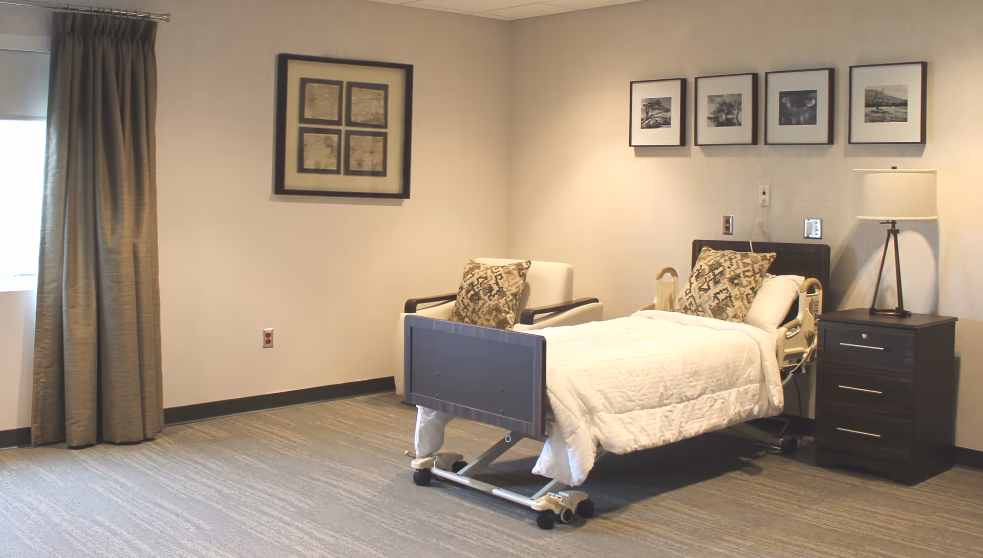 A clean and simple hospital-style room with a single adjustable bed covered with white bedding and two patterned pillows. Next to the bed is a dark wooden nightstand with a lamp on it. There is a white armchair with a patterned pillow beside the bed. The walls are light-colored and decorated with framed pictures. A window with closed curtains is on the left side of the room.