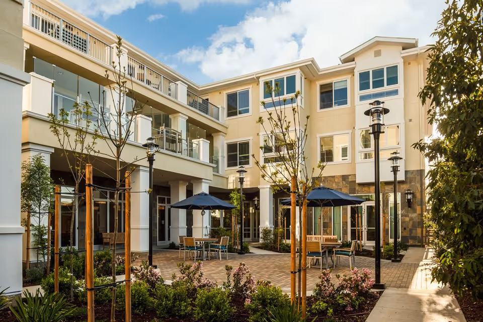 Outdoor courtyard area of a senior living facility with young trees, landscaped plants, patio tables with umbrellas, benches, and a multi-story building with balconies and large windows under a partly cloudy sky.